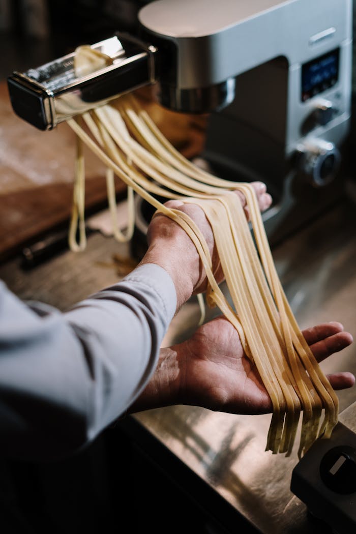 Hands guiding fresh pasta through a pasta machine in a home kitchen setting.